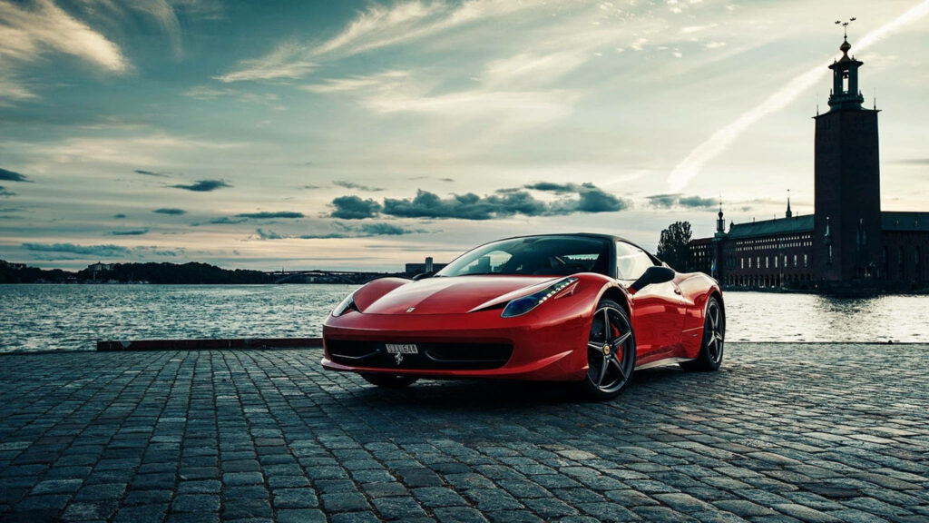 Glossy red Ferrari 458 parked by calm water at dusk, cobbled surface reflects the dying light against dramatic sky - wallpaper