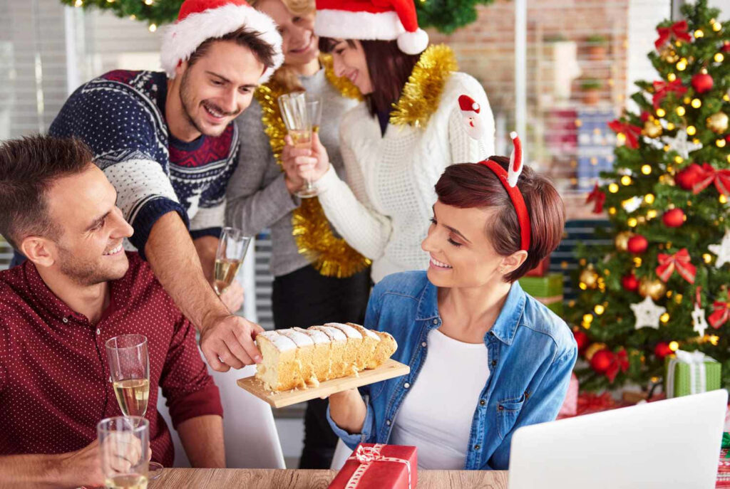 Festive Group Celebrates Holidays with Cake & Drinks by Christmas Tree Wallpaper