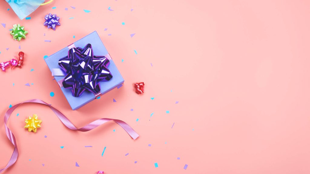 Festive flat lay purple gift box with glossy bow on soft pink background and colorful ribbons Wallpaper