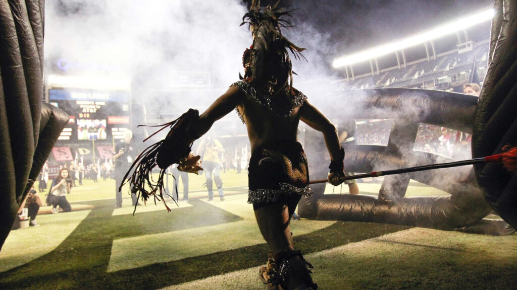 Fierce Aztec warrior in traditional attire emerging through smoke on stadium field wallpaper background