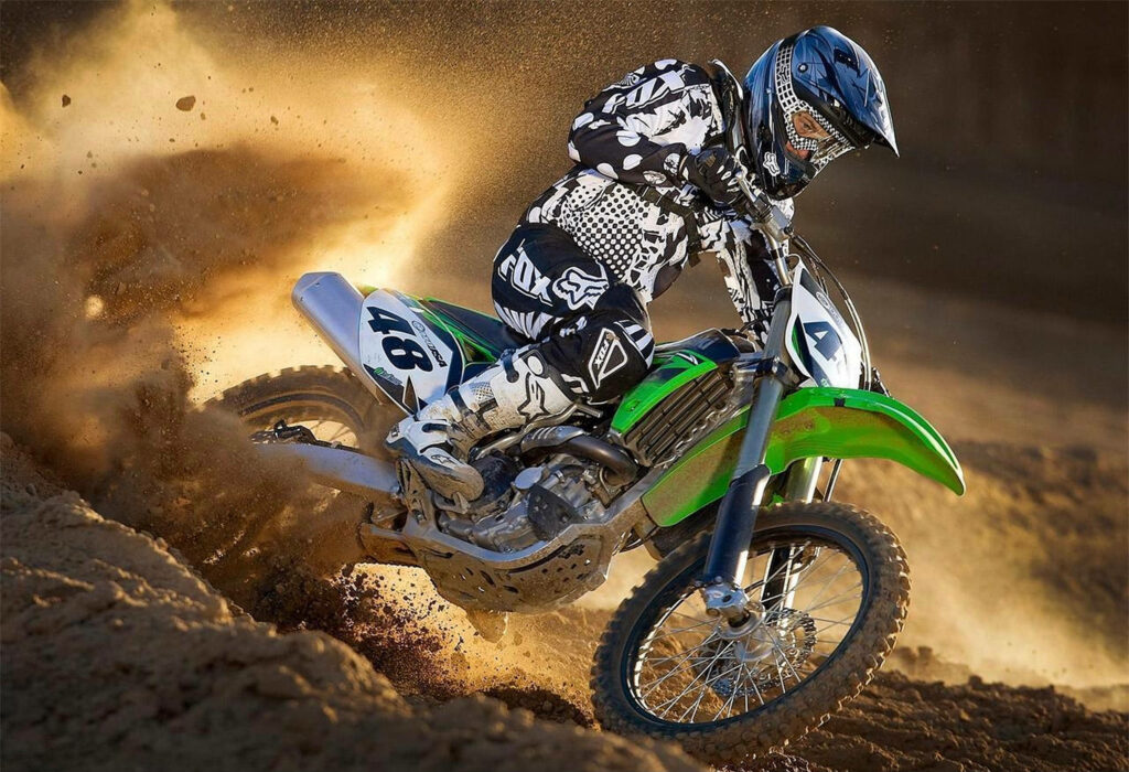 Dirt bike rider in black and white gear on green motocross bike kicking up golden sand outdoors Wallpaper