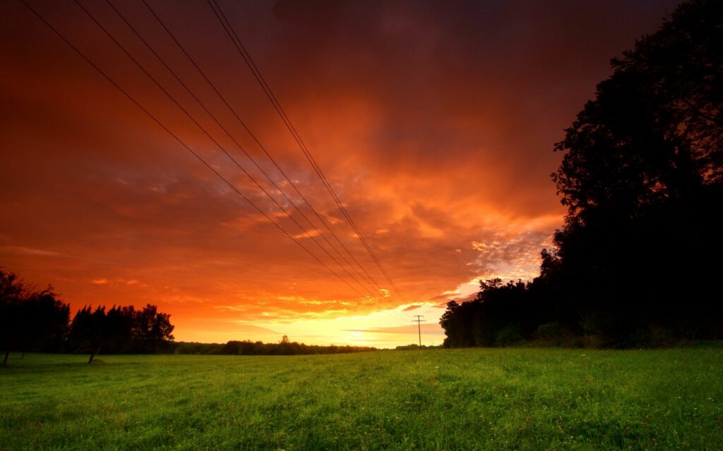 Dramatic sunset sky with fiery orange hues over green fields and silhouetted trees with power lines Wallpaper