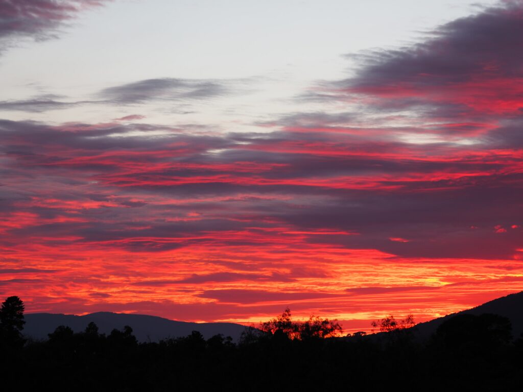 Fiery red and orange sunset over dramatic clouds with mountain and tree silhouettes wallpaper