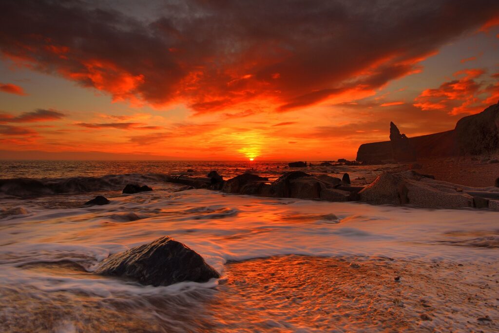 Fiery sunrise over tranquil beach with gentle waves washing dark rocks and dramatic clouds Wallpaper