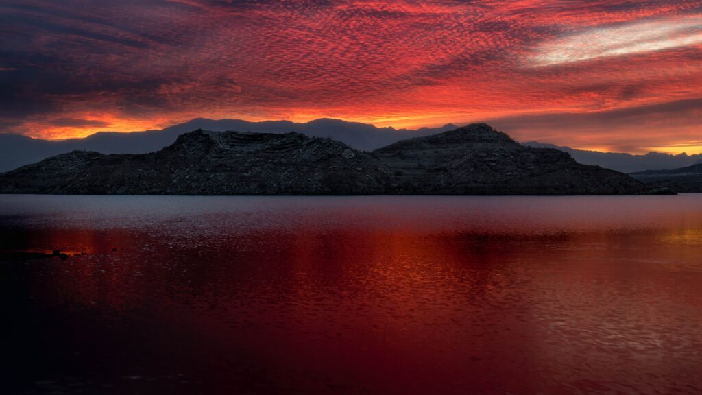 Fiery red orange sunset over Lake Mead with calm water reflecting rugged distant mountains USA Wallpaper