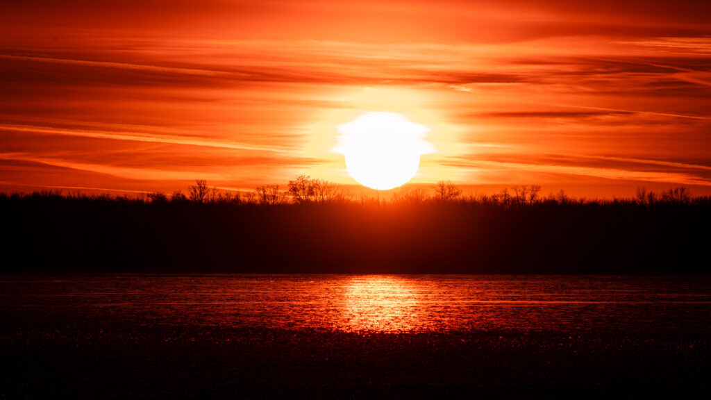 Sunset over calm water with fiery orange sky and dark tree silhouettes reflecting on surface Wallpaper
