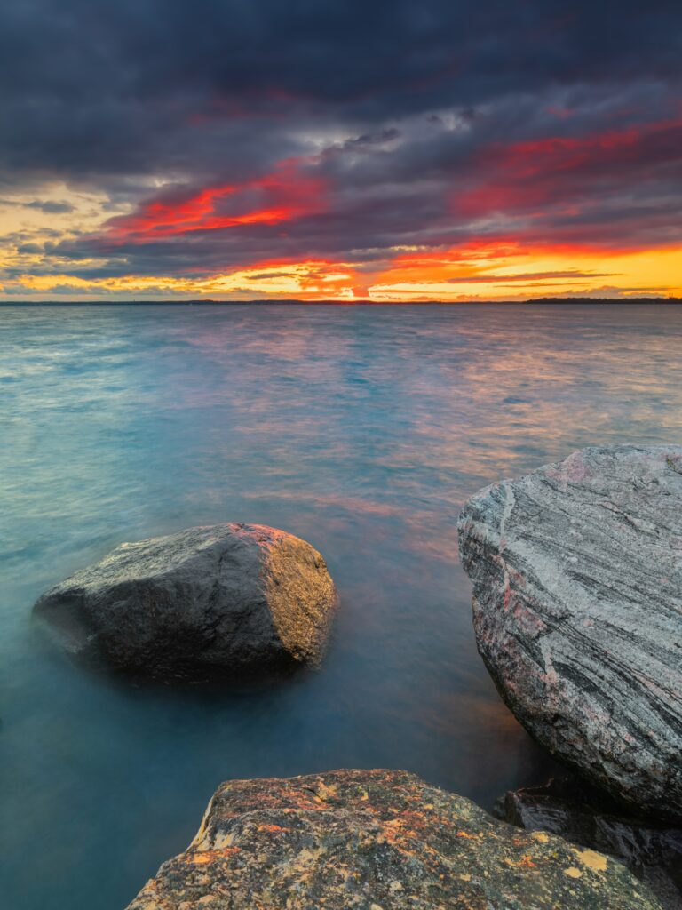 Large textured stones in calm sea at vivid fiery sunset with dramatic clouds and soft blue water Wallpaper