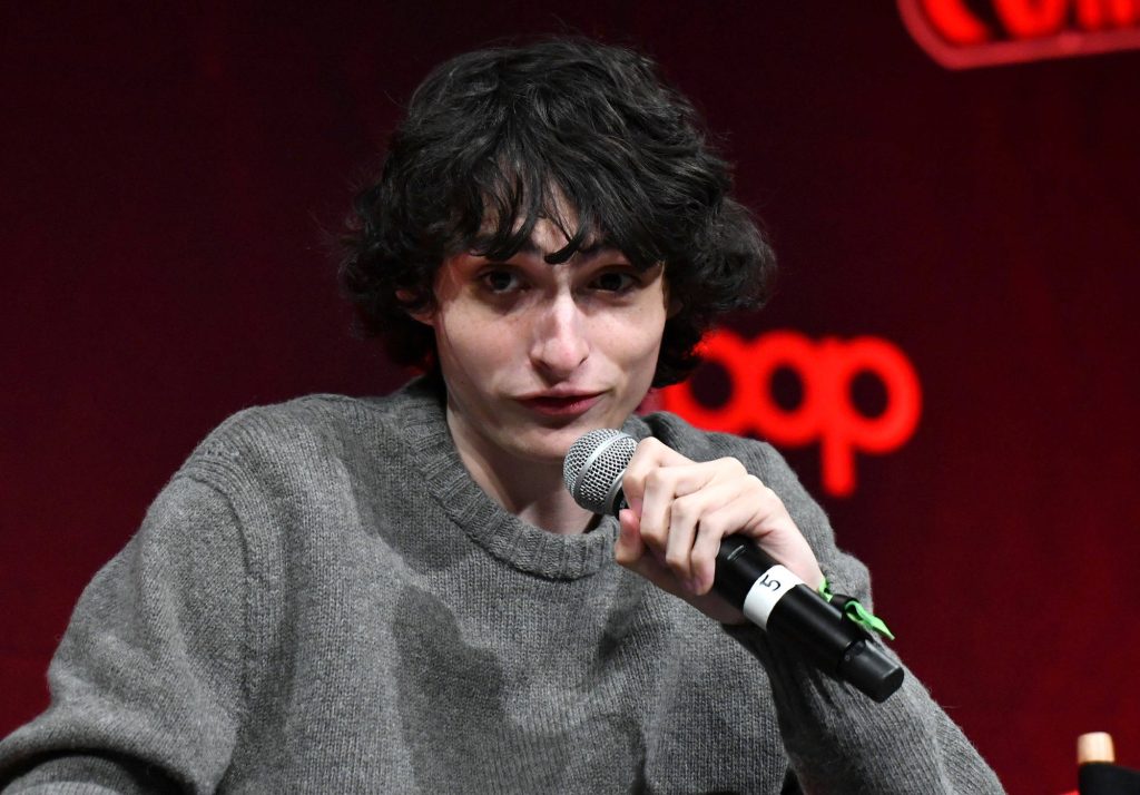 Finn Wolfhard speaking into microphone wearing gray sweater in professional interview setting Wallpaper