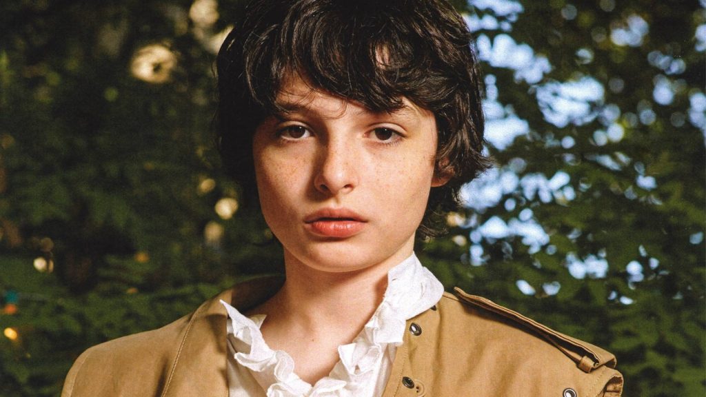 Finn Wolfhard portrait in tan coat white ruffled shirt green forest backdrop aesthetic wallpaper