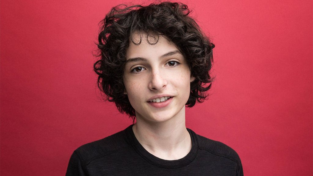 Finn Wolfhard portrait with curly hair and smile in black top red studio background wallpaper