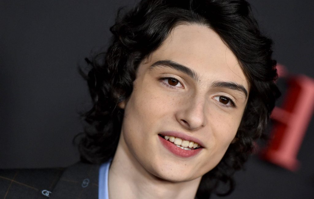 Portrait of Finn Wolfhard in a sharp suit with curly dark hair and warm smile wallpaper background