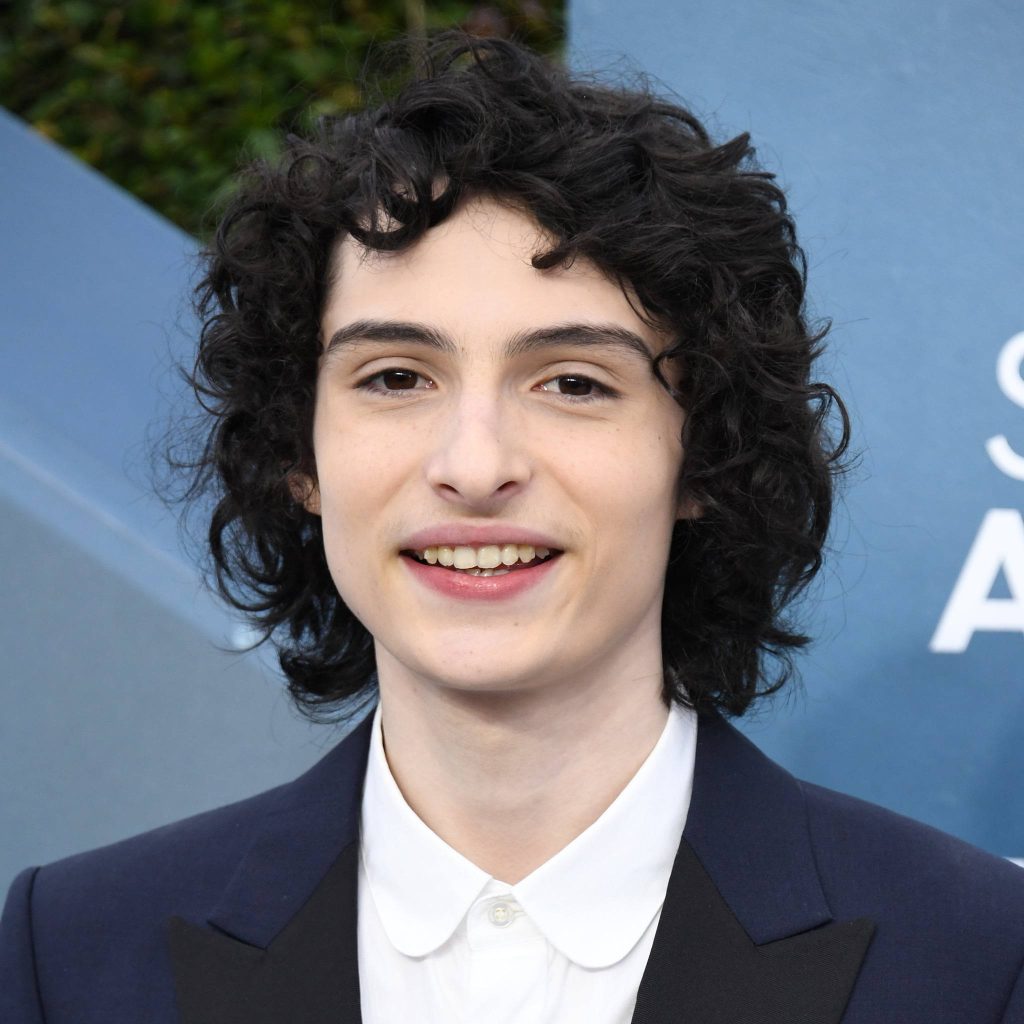 Finn Wolfhard portrait in dark suit white shirt curly hair smiling blue event backdrop wallpaper