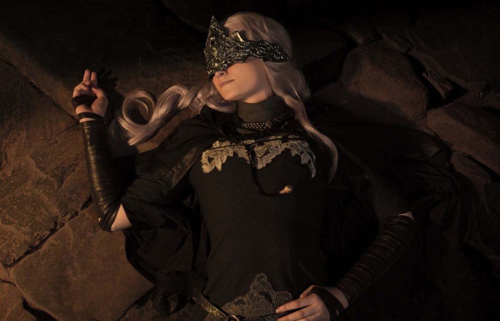 Fire Keeper cosplay from Dark Souls III sitting by bonfire in detailed dark robes and blindfold Wallpaper