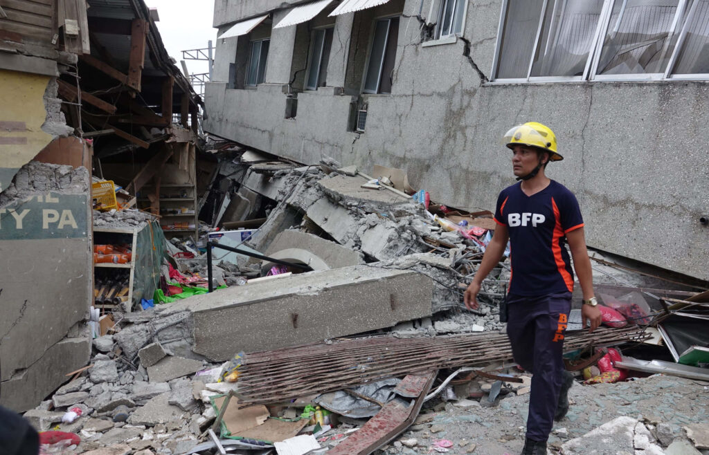Firefighter in BFP uniform walks through earthquake rubble amid collapsed buildings and debris Wallpaper