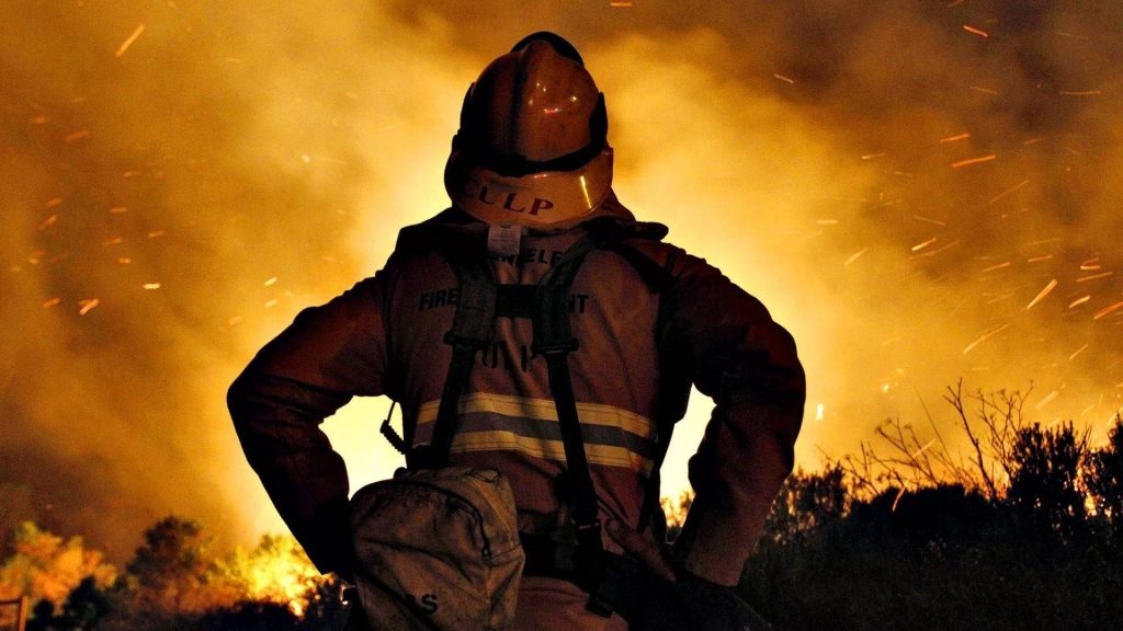 Silhouetted firefighter with CULP helmet stands amid intense blazing wildfire and smoke background Wallpaper
