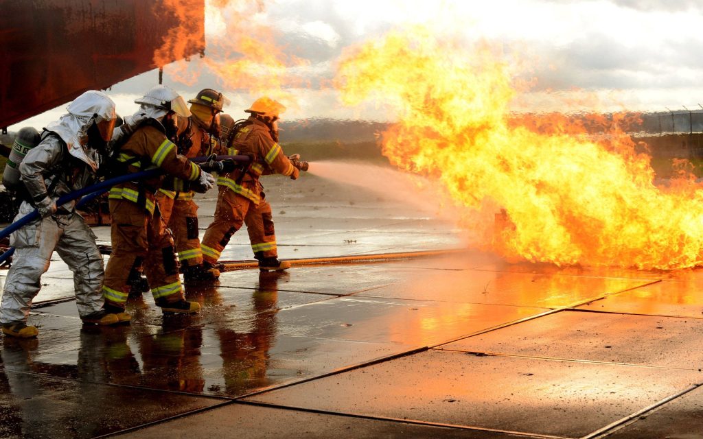 Firefighters in full gear battling intense blaze with hoses on wet tarmac during training Wallpaper