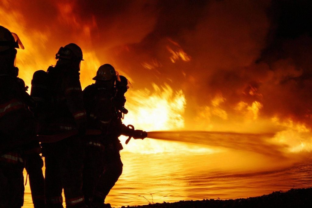 Firefighters silhouette battling massive blaze with water hose amid thick smoke and flames Wallpaper