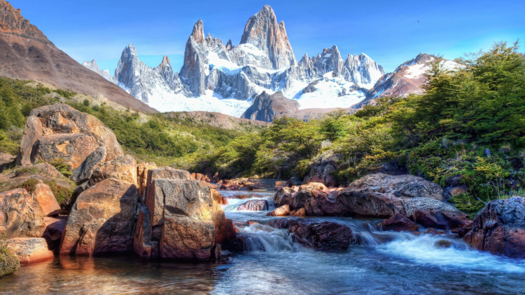 Fitz Roy peaks over clear mountain stream with colorful rocks and lush green trees panorama Wallpaper