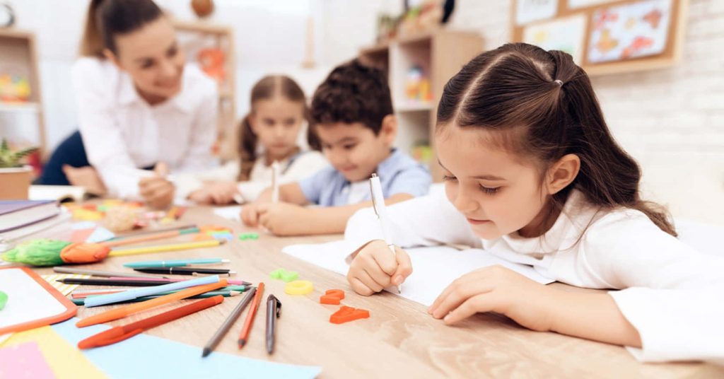 Focused children learning in classroom with girl writing colorful pens and teacher supervising Wallpaper
