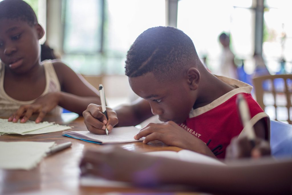 Children studying focused in bright classroom boy in red shirt writing in notebook academic dedication Wallpaper