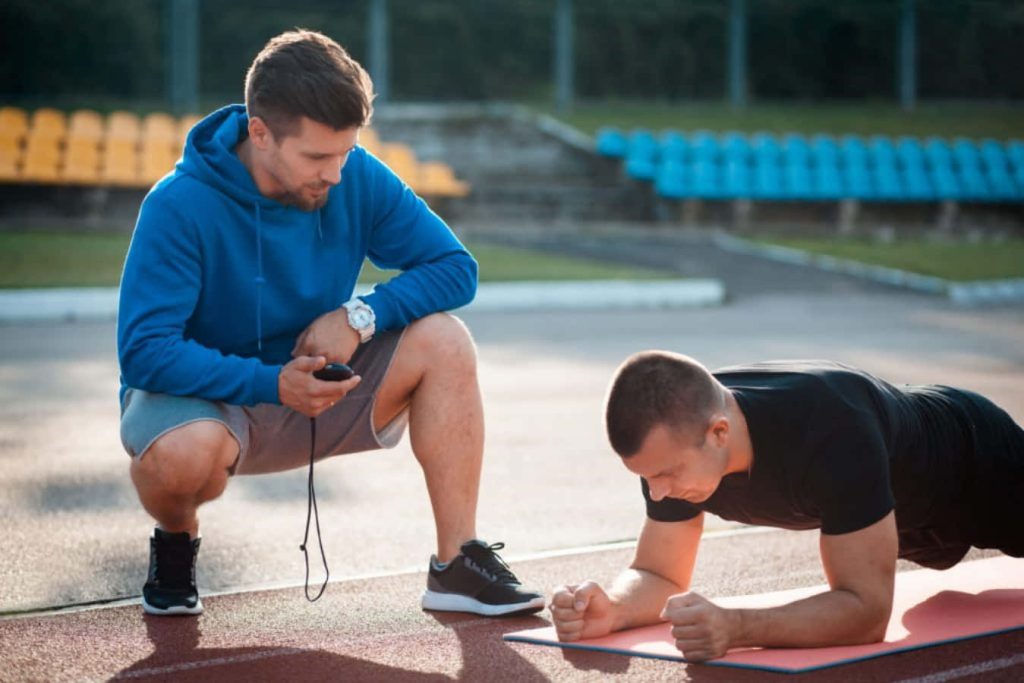 Personal trainer in blue hoodie coaching athlete doing plank on red running track fitness wallpaper