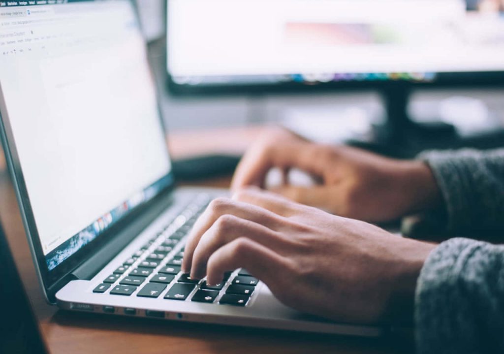 Close-up of hands typing on laptop keyboard with blurred monitor background workspace wallpaper