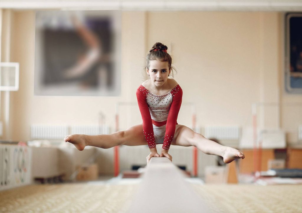 Young gymnast in red white leotard doing straddle split on balance beam in training gym Wallpaper