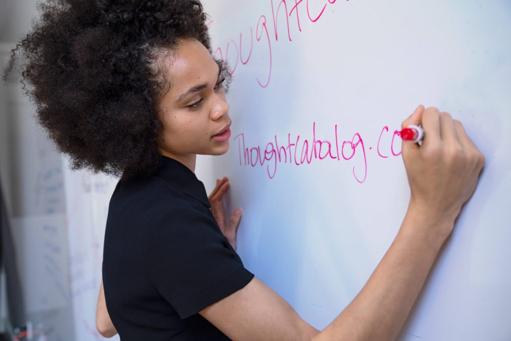 Young woman with curly hair writing Thoughtcatalog com on whiteboard in bright workspace Wallpaper