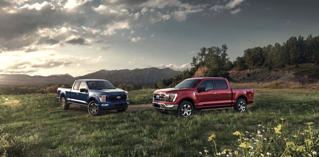 Two Ford F-150 trucks parked in a scenic mountain field with cloudy sky and trees background Wallpaper