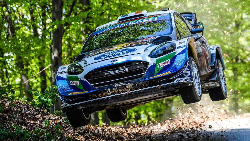 Ford Fiesta rally car airborne over forest dirt trail with lush greenery and autumn leaves wallpaper