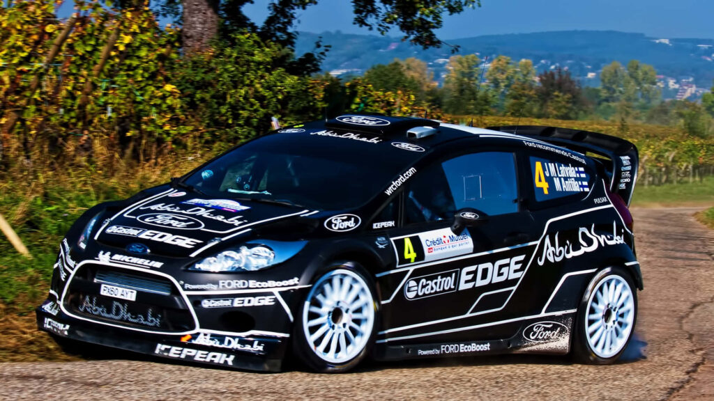 Ford Fiesta rally car speeding on scenic countryside road with vibrant sponsorship decals wallpaper