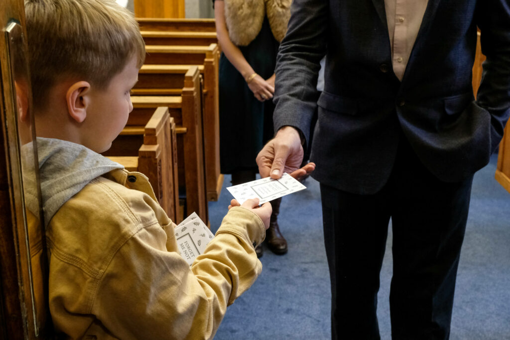 Remembrance Blooms: A Tender Moment at a Funeral as a Young Boy Distributes Forget-Me-Not Cards amid a Floral Backdrop Wallpaper