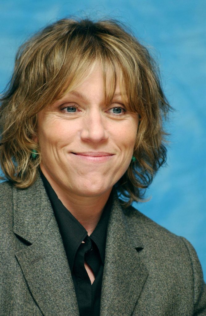 Frances McDormand portrait smiling in grey blazer black shirt teal earrings blue background Wallpaper