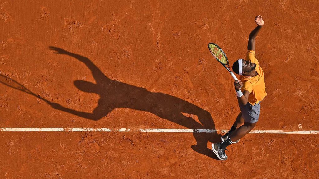 Tennis star Frances Tiafoe powerful serve red clay court dynamic action shot wallpaper