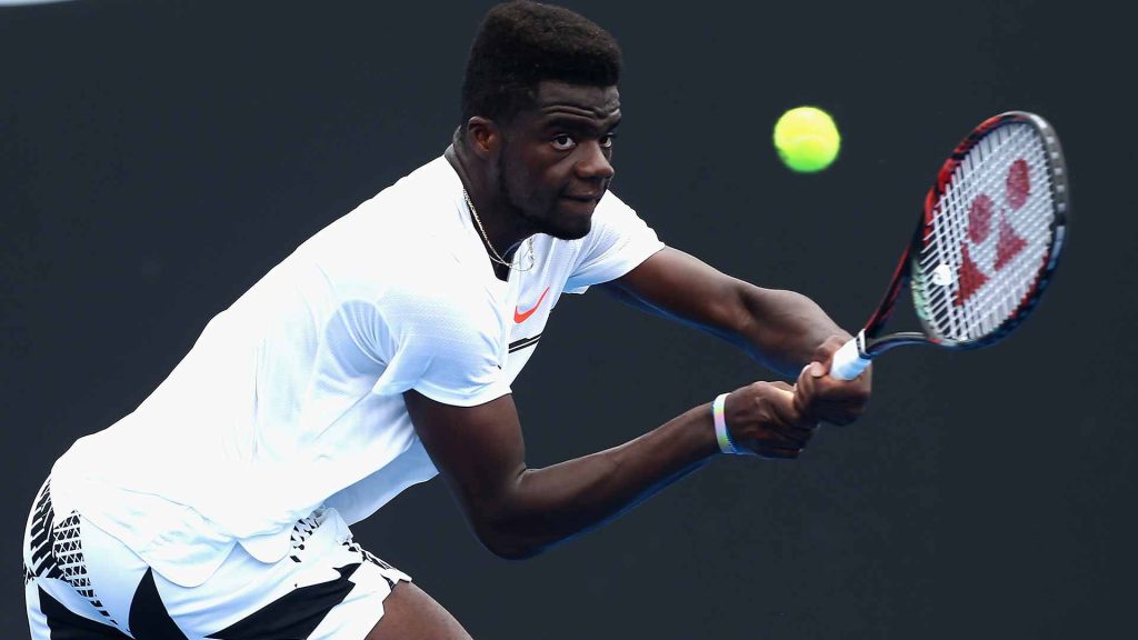Frances Tiafoe hitting powerful two-handed backhand in white kit tennis wallpaper background