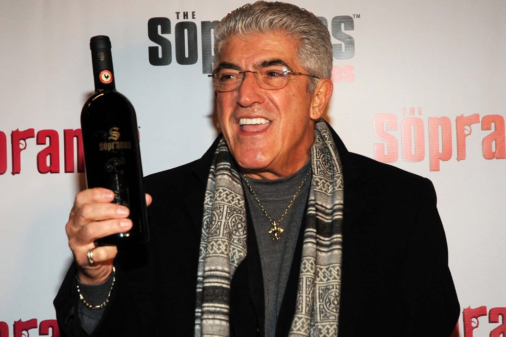 Frank Vincent smiling with Sopranos wine bottle wearing dark coat turtleneck and scarf wallpaper