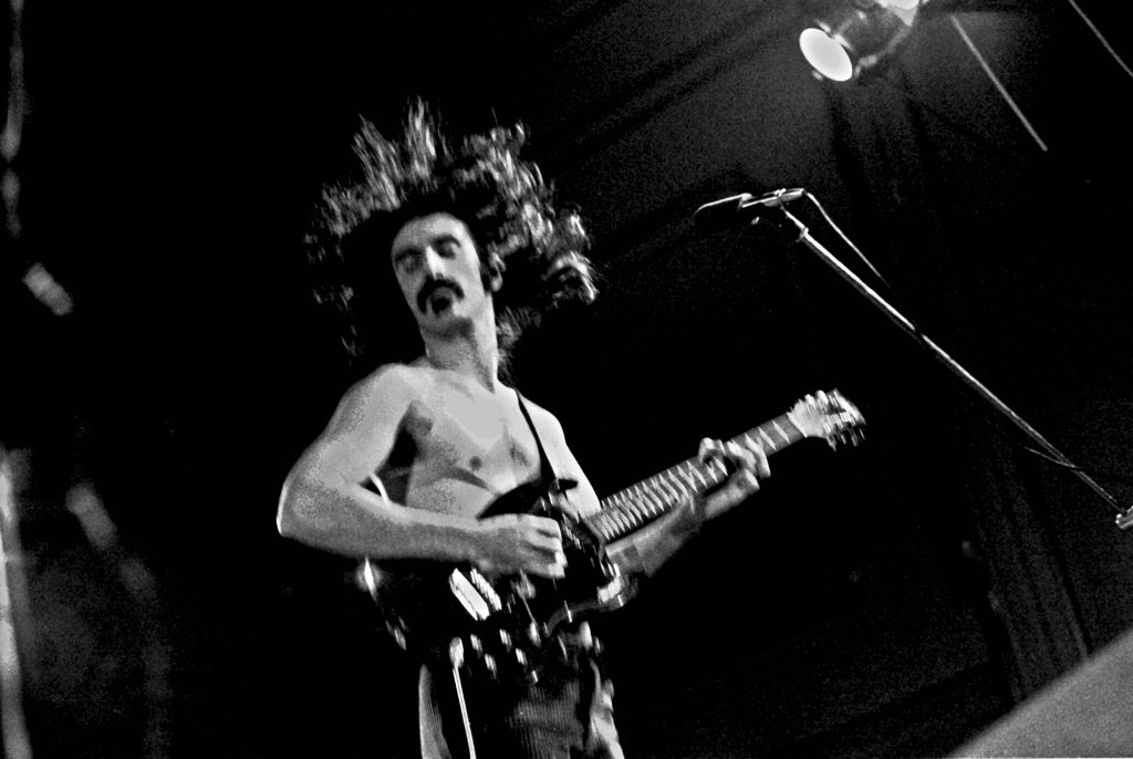 Frank Zappa shirtless playing electric guitar on stage in dramatic black and white concert photo Wallpaper