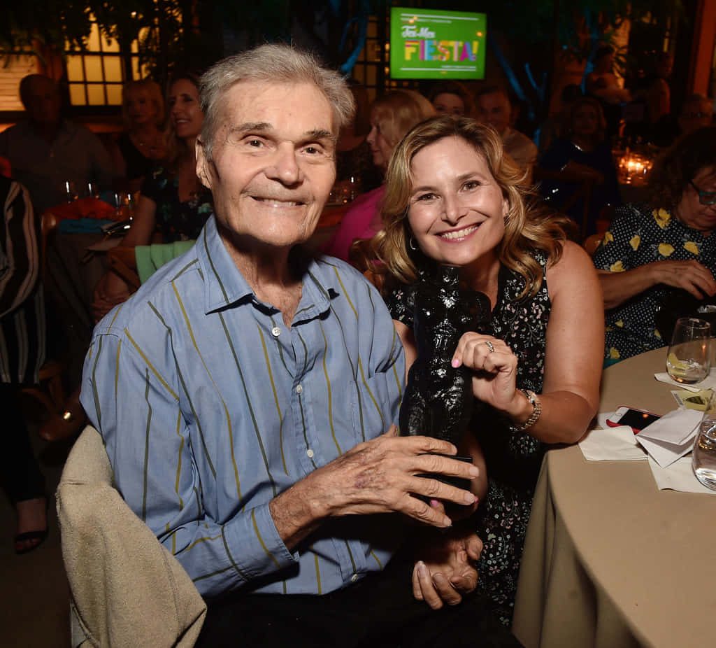 Fred Willard smiling with companion holding black trophy at intimate banquet in warm lighting Wallpaper