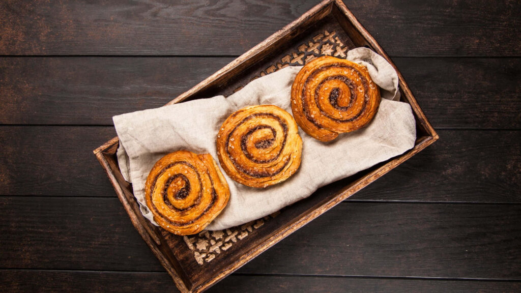 Three golden brown cinnamon rolls on rustic linen cloth wooden tray cozy kitchen wallpaper