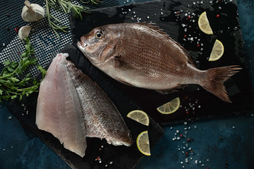 Fresh whole bream fish fillets lemon garlic herbs salt peppercorns on dark slate surface Wallpaper