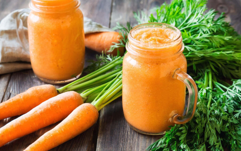 Freshly blended carrot juice in glass jars with vibrant carrots and green tops on wood table Wallpaper