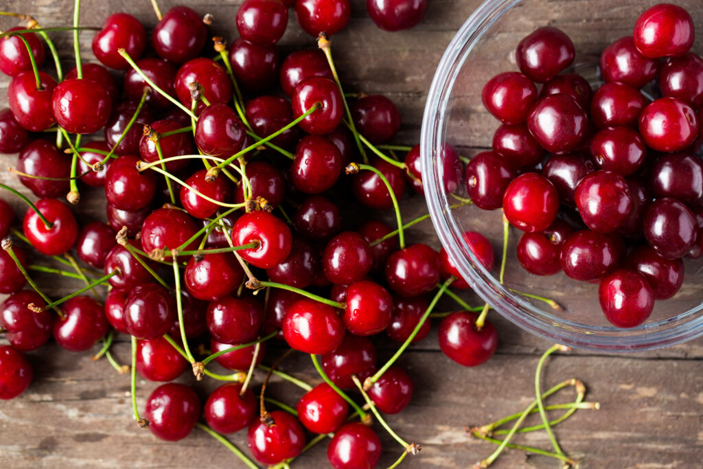 Fresh red cherries with green stems on rustic wood next to a clear bowl summer wallpaper background