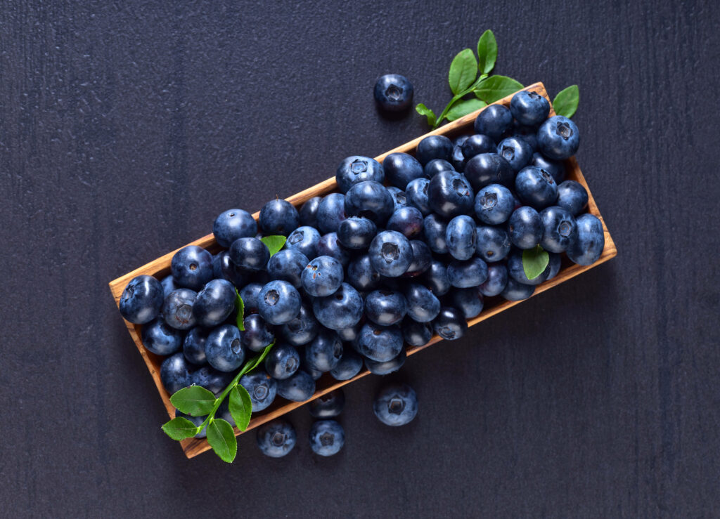 Rectangular wooden tray with fresh ripe blueberries and green leaves on dark textured surface Wallpaper