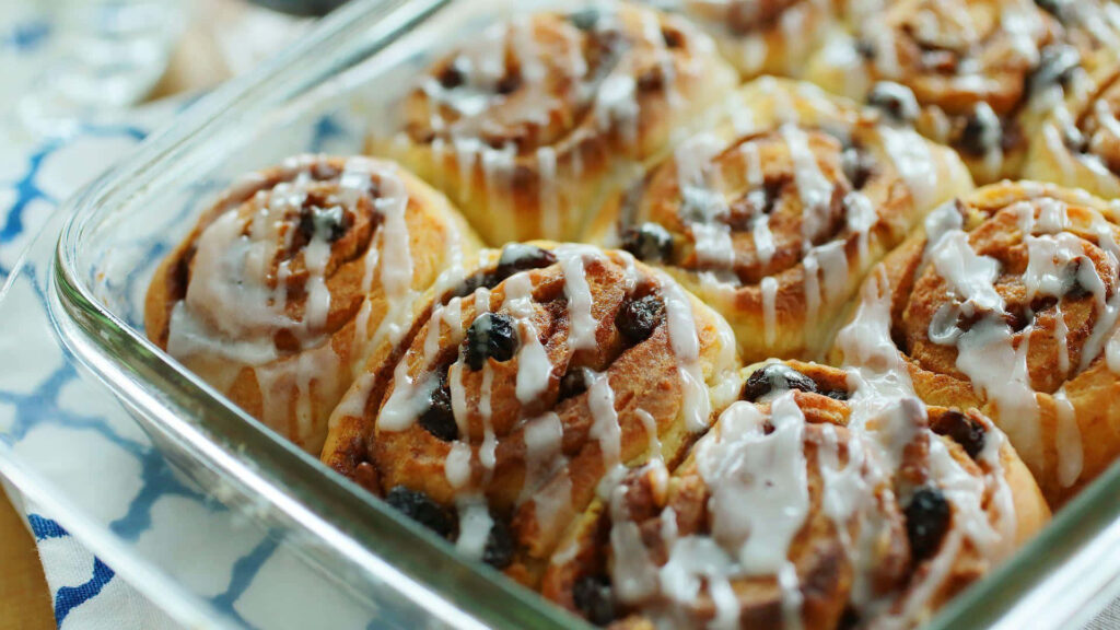 Freshly baked golden cinnamon rolls with raisins and creamy icing on bakery wallpaper background
