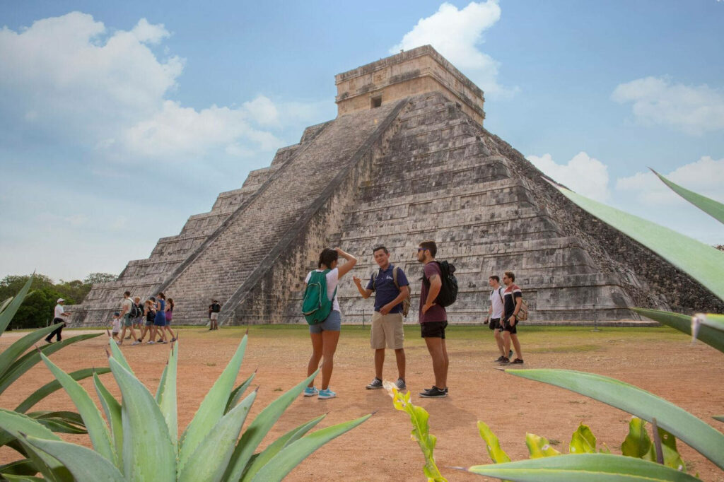 Friends exploring ancient Chichen Itza pyramid under blue sky with lush green plants nearby Wallpaper