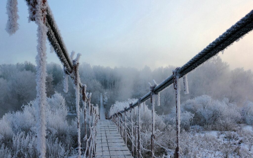Frost-covered narrow rope bridge over icy winter landscape with fog and frozen trees wallpaper