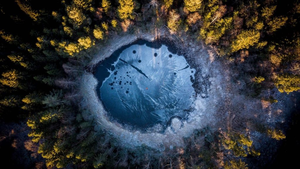 Aerial view of frozen circular pond in golden forest with lone person in red winter scene wallpaper