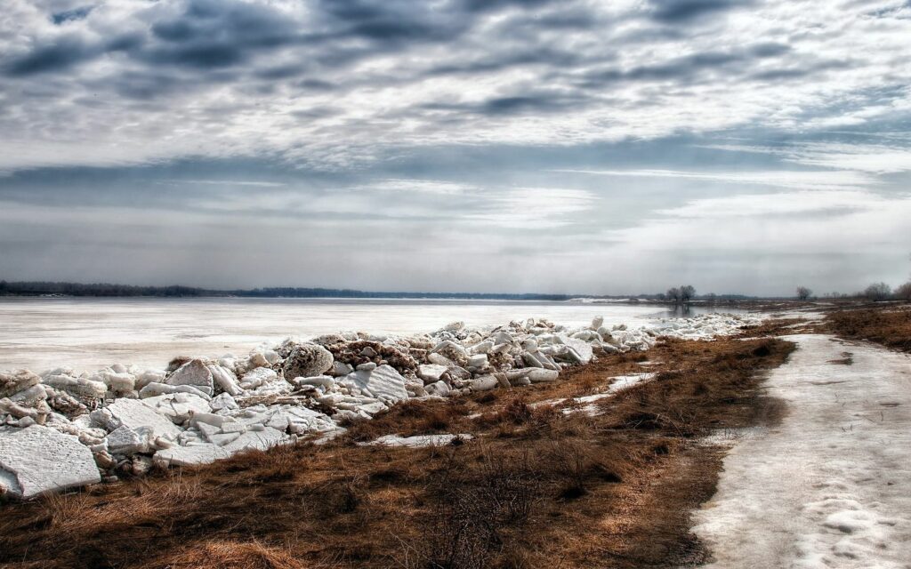 Frozen riverbank with jagged ice sheets under dramatic cloudy sky in winter landscape wallpaper