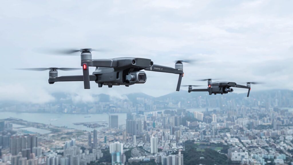 Two advanced drones with metallic designs flying over a modern cityscape under an overcast sky Wallpaper