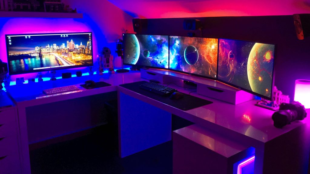 Futuristic gaming room with L-shaped white desk triple galaxy monitors purple blue lighting R2-D2 figure Wallpaper